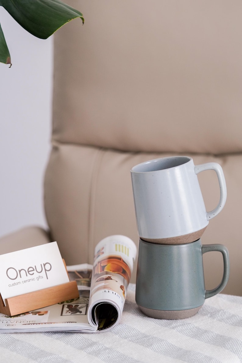 May include: Two ceramic mugs, one light blue and one teal, stacked on top of each other. The mugs have a two-tone design with a brown base. A magazine and a wooden business card holder with the text "Oneup custom ceramic gifts" are in the foreground.