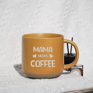 May include: Mustard yellow ceramic coffee mug with the text "MAMA NEEDS COFFEE" in white. The mug has a gray base and a curved handle. The mug is on a white textured surface.