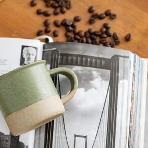 May include: A handmade ceramic mug with a green speckled upper half and a tan lower half. The mug has a curved handle and sits on a stack of books with a black and white image. Coffee beans are scattered around the mug.