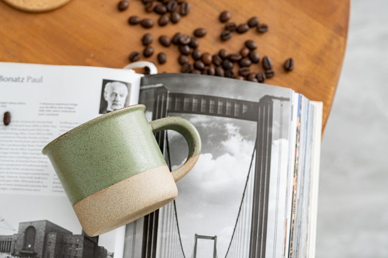 May include: A handmade ceramic mug with a green speckled upper half and a tan lower half. The mug has a curved handle and sits on a stack of books with a black and white image. Coffee beans are scattered around the mug.