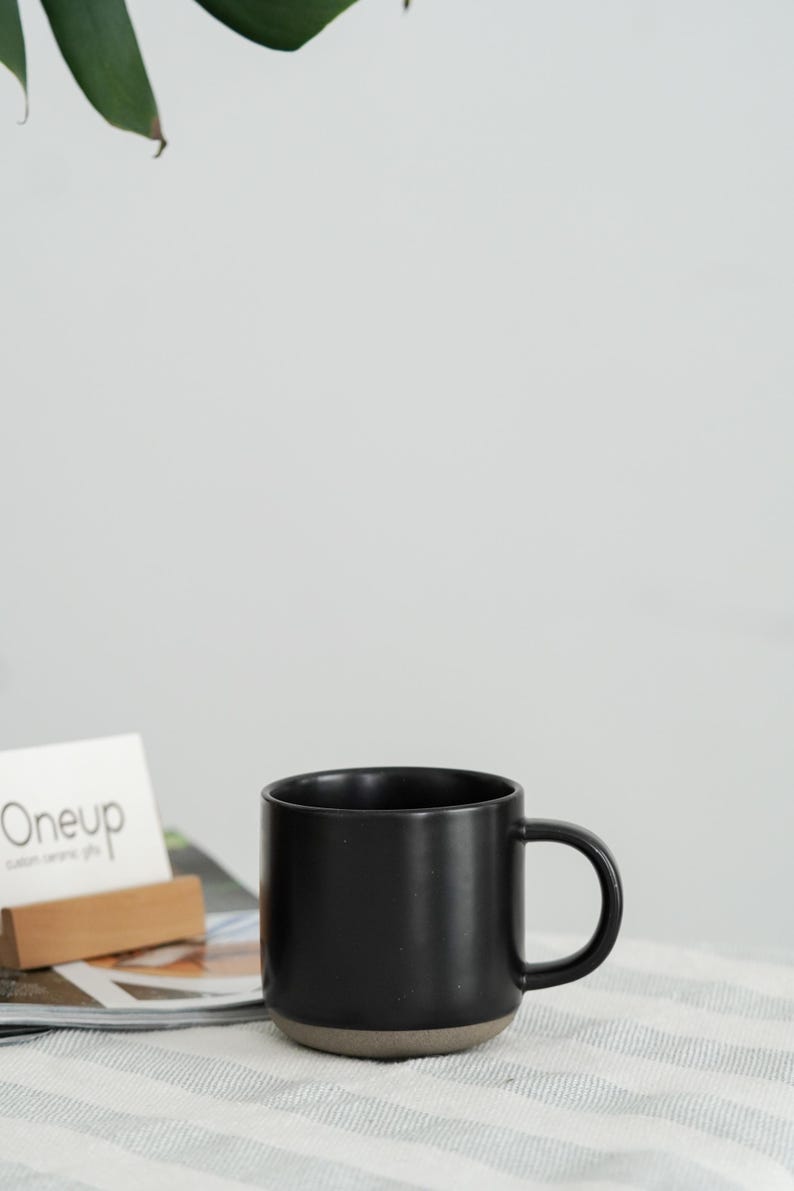 Pode incluir: Uma caneca de cer&acirc;mica preta com base cinza escuro e uma al&ccedil;a arredondada. A caneca est&aacute; sobre uma superf&iacute;cie listrada ao lado de uma revista e um porta-cart&otilde;es de madeira com a palavra "Oneup" impressa em um cart&atilde;o.
