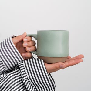 May include: A person's hand holding a light green ceramic mug with a brown rim. The mug has a speckled texture.