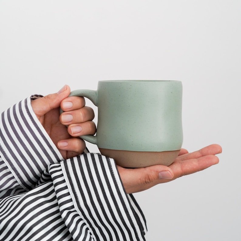 May include: A person's hand holding a light green ceramic mug with a brown rim. The mug has a speckled texture.