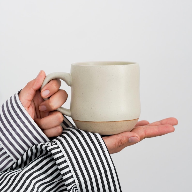 May include: A person holding a light beige ceramic mug with a rounded bottom and a slightly flared rim. The mug is being held by a hand wearing a black and white striped shirt.