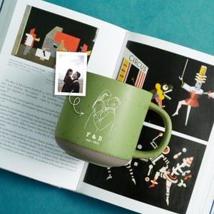 May include: Green ceramic mug with a white line drawing of a couple embracing. The mug has a speckled finish and a grey base. The text "F & B est. 2015" is below the drawing. The mug is on a book.