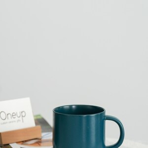 Pode incluir: Uma caneca de cer&acirc;mica azul-petr&oacute;leo com base cinza e uma al&ccedil;a arredondada. A caneca est&aacute; sobre uma superf&iacute;cie listrada. Um cart&atilde;o de visita com a palavra "Oneup" est&aacute; no fundo.