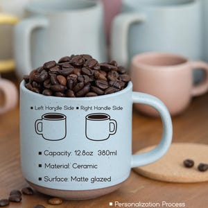 May include: A light blue ceramic mug with a matte glaze and a 12.8 ounce capacity. The mug has a handle on the right side and features a black outline of a mug on each side. The mug is filled with coffee beans and is sitting on a wooden surface. The text on the mug reads "Left Handle Side Right Handle Side" and "Capacity: 12.8oz 380ml". The text below the mug reads "Food safe", "Microwave safe", and "Dishwasher safe". The text on the right side of the mug reads "Personalization Process", "Laser Engraving", and "Fill with colored ceramic paint (non-toxic)".