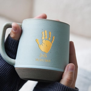 Op de afbeelding: Een lichtblauwe keramische mok met een bruine basis en handvat. De mok heeft een gouden handafdruk en de naam "Stella" met de datum "10/09/2015" in goud gedrukt. De mok wordt door een persoon vastgehouden.