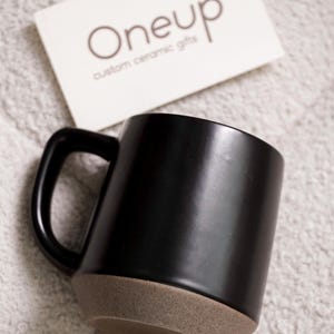 May include: A black ceramic mug with a brown base and a black handle. A white card with the text "Oneup custom ceramic gifts" is in the background. The mug is a simple, modern design.