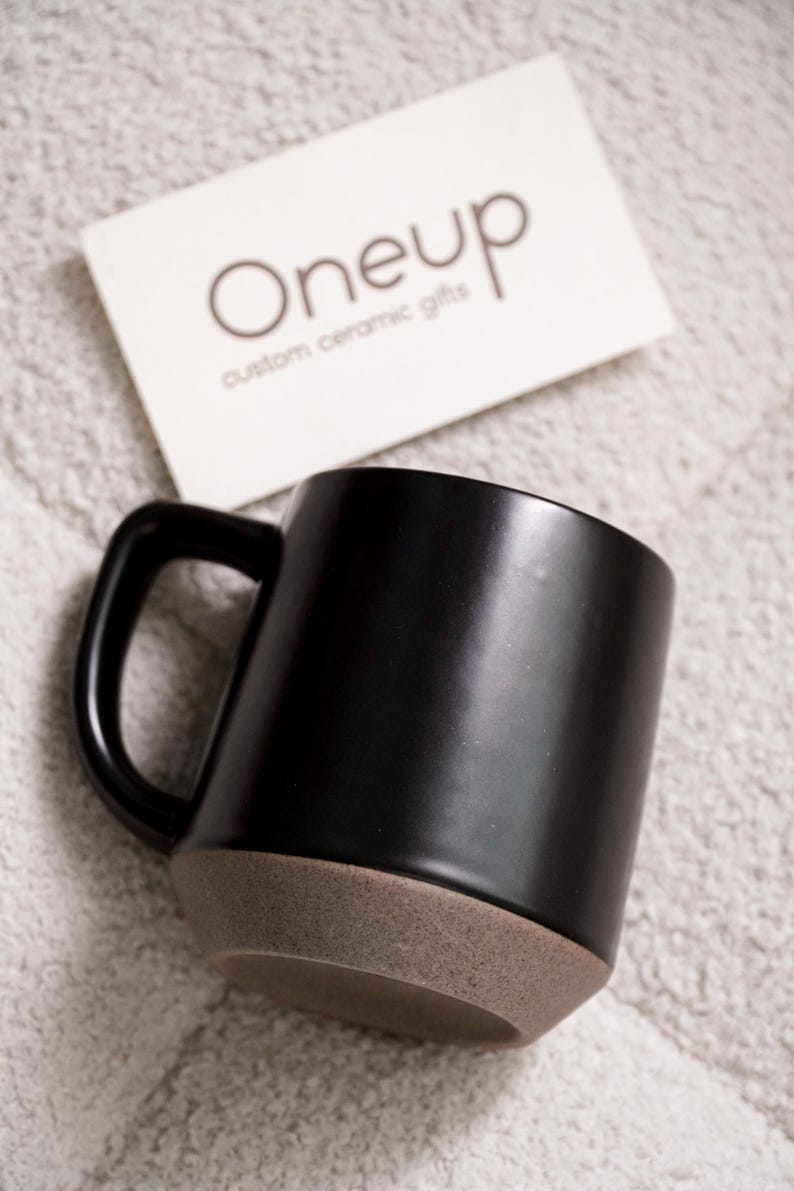 May include: A black ceramic mug with a brown base and a black handle. A white card with the text "Oneup custom ceramic gifts" is in the background. The mug is a simple, modern design.