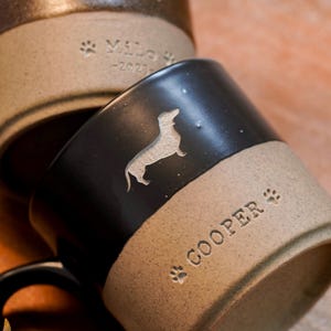 May include: Two ceramic mugs with a two-tone design featuring a dark upper section and a speckled light brown lower section. The front mug displays a silhouette of a dachshund and the name COOPER with paw prints. The back mug shows a cat silhouette.