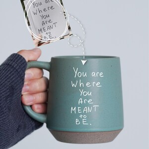 Könnte beinhalten: Eine türkisfarbene Keramiktasse mit braunem Boden, auf der handschriftlich der Satz "You are Where You Are MEANT to BE" steht. Ein kleiner Notizblock mit demselben Text wird über der Tasse gehalten.