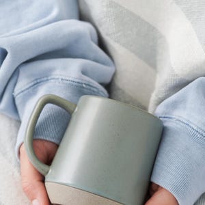 May include: A gray ceramic mug with a light tan base is held in both hands. The mug has a simple, modern design with a curved handle. The person holding the mug is wearing a light blue sweatshirt.