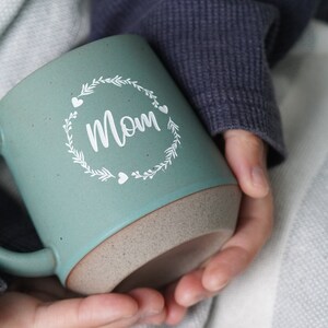 May include: A teal ceramic mug with a white design of a wreath with the word "Mom" inside. The mug has a speckled brown rim.
