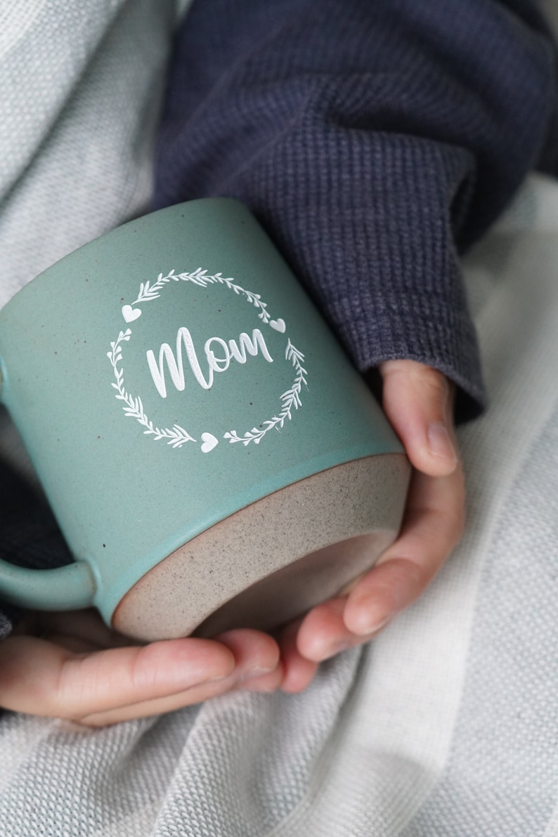 May include: A teal ceramic mug with a white design of a wreath with the word "Mom" inside. The mug has a speckled brown rim.