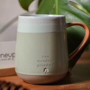 Może przedstawiać: Ceramiczny kubek z dwukolorowym wykończeniem, posiadający białą górną część i nakrapiany jasnozielony korpus. Napis "tree mendous grandma" jest wytłoczony z przodu nad małą ikoną drzewa. Posiada zakrzywiony, gładki brązowy uchwyt.