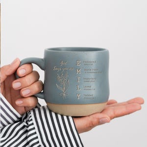 Puede incluir: Una taza de cerámica azul mate con acabado moteado y base de color tostado natural. La taza presenta una ilustración floral y el texto: God Says you are E Embraced, M Made New, I Important, L Loved, Y Yielded, junto a versículos bíblicos.