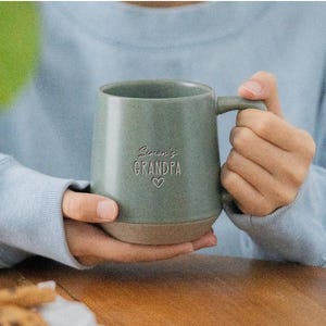 Puede incluir: Una taza de cerámica verde salvia con un acabado moteado y una base de color tostado en contraste. La parte frontal de la taza presenta el texto grabado "Simon's GRANDPA" sobre un pequeño símbolo de corazón. Tiene una asa y forma redondeada.