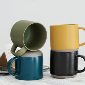 Pode incluir: Quatro canecas de cer&acirc;mica empilhadas em tons de verde, verde-azulado, amarelo e preto. Cada caneca tem uma base cinza contrastante e uma al&ccedil;a curva. As canecas est&atilde;o sobre uma superf&iacute;cie branca com uma revista e um objeto de madeira ao fundo.