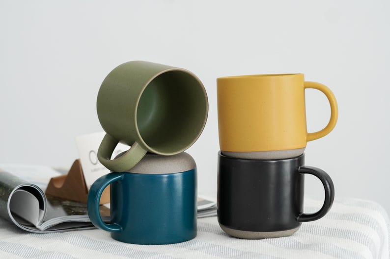 Pode incluir: Quatro canecas de cer&acirc;mica empilhadas em tons de verde, verde-azulado, amarelo e preto. Cada caneca tem uma base cinza contrastante e uma al&ccedil;a curva. As canecas est&atilde;o sobre uma superf&iacute;cie branca com uma revista e um objeto de madeira ao fundo.