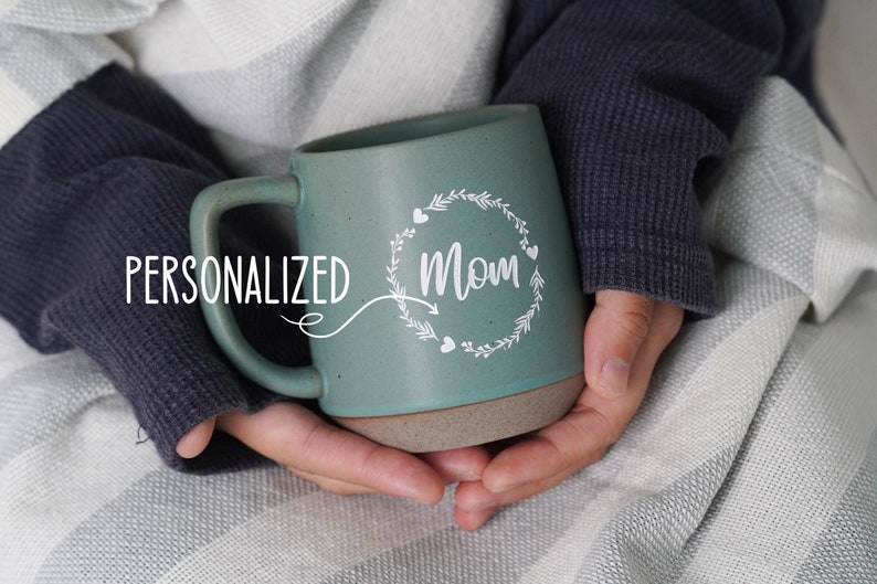 May include: A personalized teal ceramic mug with a white design of a wreath with the word "Mom" in the center. The mug has a brown rim and base.