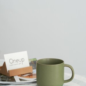 Pode incluir: Uma caneca de cer&acirc;mica verde com uma al&ccedil;a, sobre uma superf&iacute;cie listrada. A caneca tem uma textura salpicada e uma base cinza escuro. Um porta-cart&otilde;es com a inscri&ccedil;&atilde;o "Oneup" est&aacute; no fundo.