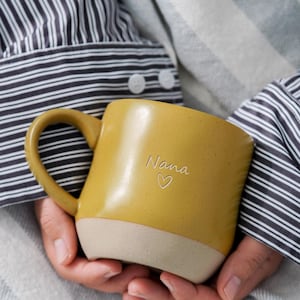 May include: A mustard yellow ceramic mug with a speckled texture and a smooth, light beige base. The mug features a curved handle and the word Nana written in a delicate script font above a small, simple outline of a heart on the front surface.