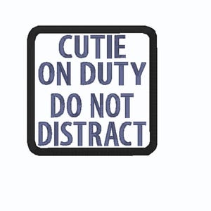 May include: A square sign with a black border and white background. The sign features the words "CUTIE ON DUTY DO NOT DISTRACT" in blue, stylized text. The sign is centered on a white background.