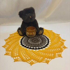 May include: A brown teddy bear sits atop a yellow and brown crocheted doily, with a small honey pot. The doily is designed to resemble a sunflower. The honey pot has the word "Honey" written on it.