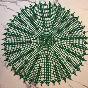 May include: A handmade, circular, green crochet doily. The doily features a detailed openwork design with a central spiral pattern and a scalloped edge. The color is a deep, vibrant green, and the doily is displayed on a marble-patterned surface.