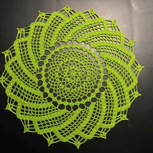 May include: A lime green, crocheted doily with a spiral design. The handmade piece features an open, lacy pattern and a scalloped edge. The circular textile is displayed on a dark surface.