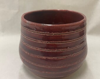 May include: A deep red ceramic planter with a rounded shape and horizontal lines. The planter is glazed and has a smooth finish.