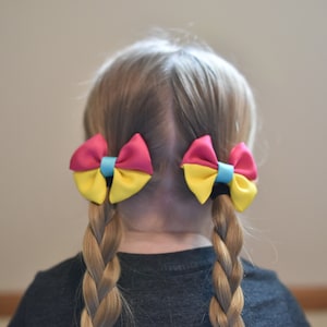 May include: A child's blonde hair styled in two braids, adorned with vibrant hair bows. Each bow features a combination of pink, yellow, and blue, creating a colorful accessory. The bows are secured with black hair ties.
