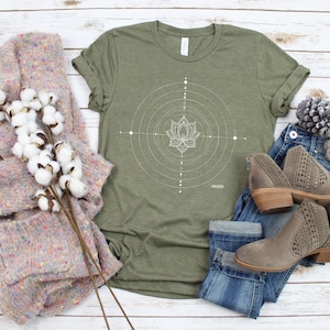 Pode incluir: Uma camiseta verde com um design de flor de l&oacute;tus geom&eacute;trico branco e a palavra "HAGEN" impressa na parte inferior. A camiseta est&aacute; colocada sobre uma superf&iacute;cie de madeira branca com um su&eacute;ter rosa, jeans azul e botas marrons.