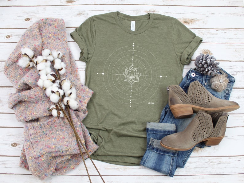 Pode incluir: Uma camiseta verde com um design de flor de l&oacute;tus geom&eacute;trico branco e a palavra "HAGEN" impressa na parte inferior. A camiseta est&aacute; colocada sobre uma superf&iacute;cie de madeira branca com um su&eacute;ter rosa, jeans azul e botas marrons.