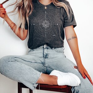 Pode incluir: Uma camiseta cinza escura com um design geom&eacute;trico branco com uma flor de l&oacute;tus. A camiseta tem a palavra "HUSTLE" impressa na parte inferior.
