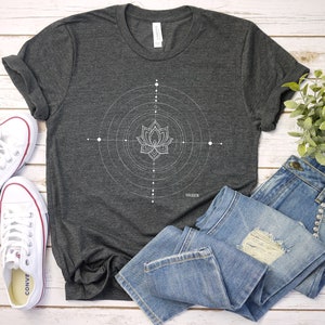 Pode incluir: Uma camiseta cinza escura com um design geom&eacute;trico branco com uma flor de l&oacute;tus. A camiseta &eacute; combinada com jeans azul e t&ecirc;nis brancos.