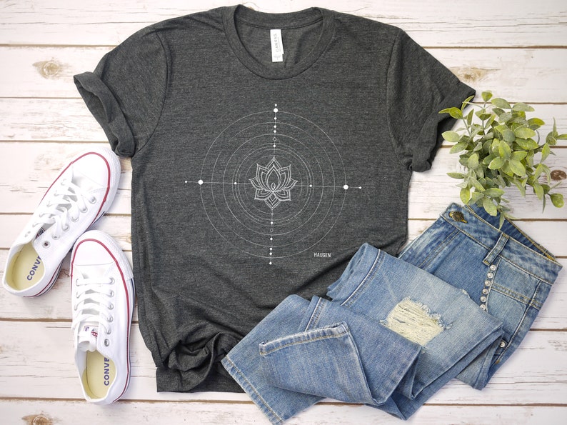 Pode incluir: Uma camiseta cinza escura com um design geom&eacute;trico branco com uma flor de l&oacute;tus. A camiseta &eacute; combinada com jeans azul e t&ecirc;nis brancos.