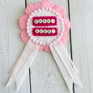 May include: A circular felt award ribbon featuring a scalloped light pink base, a white center with a pom-pom trim, and two dark pink felt strips. The text DRAMA QUEEN is spelled out in white letter beads with gold lettering on the dark pink strips.