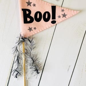 May include: A pink felt flag with the word "Boo!" in black, decorated with silver glitter stars. The flag is attached to a wooden dowel with silver and black fringe, ideal for Halloween decorations.