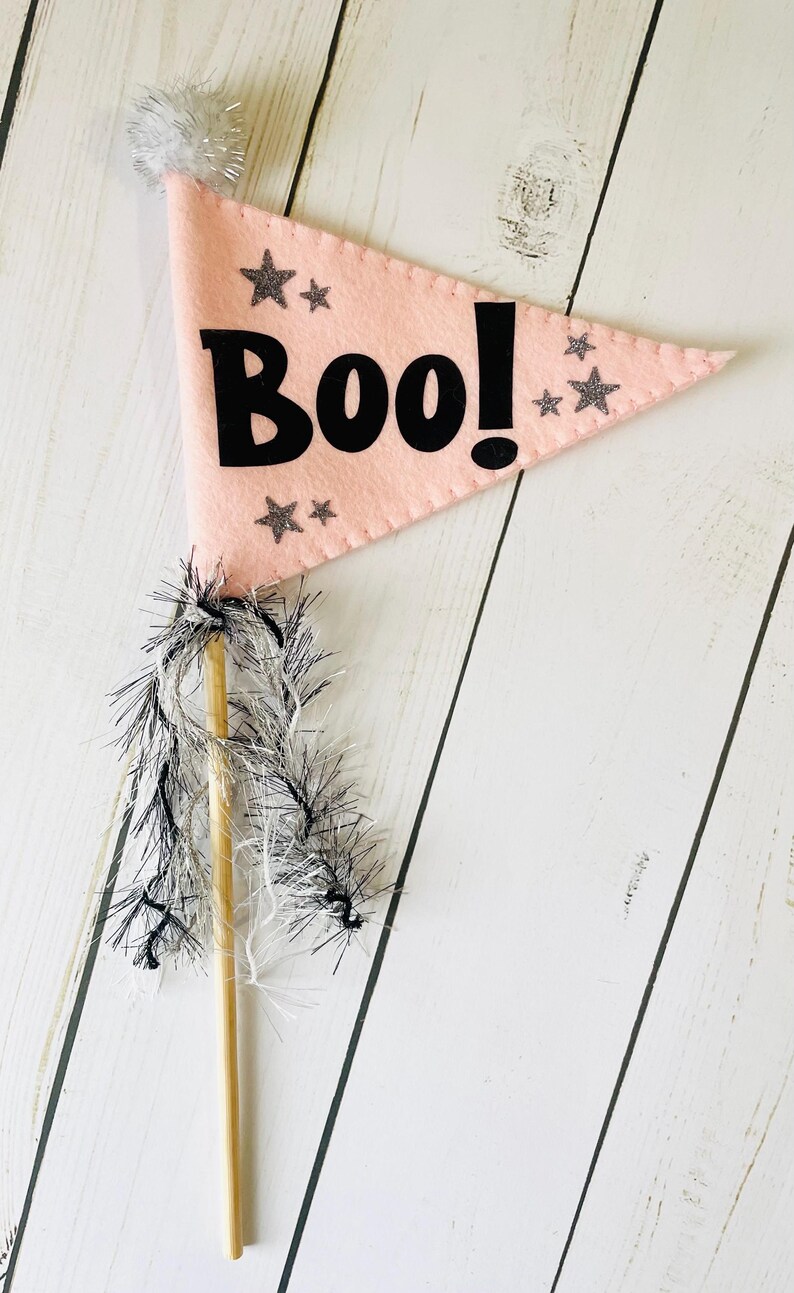 May include: A pink felt flag with the word "Boo!" in black, decorated with silver glitter stars. The flag is attached to a wooden dowel with silver and black fringe, ideal for Halloween decorations.