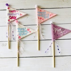 May include: Three felt pennants with wooden sticks. The pennants are decorated with different messages for Mother's Day. One says "Happy Mother's Day" in pink and white lettering with pink flowers. Another says "Best Mum Ever" in blue and white lettering with blue flowers. The last one says "Mom" in white lettering with a pink heart.