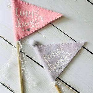 May include: Two felt pennants with wooden sticks and white string. The pennants are pink and purple and have the text "tiny dancer" written in white on them.