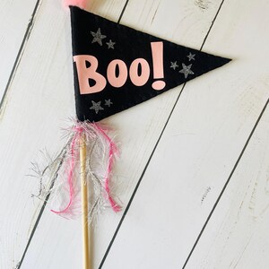 May include: A black felt flag with the word "Boo!" in pink, decorated with silver glitter stars. The flag has a pink pom-pom and is attached to a wooden dowel with pink and silver fringe, ideal for Halloween celebrations.