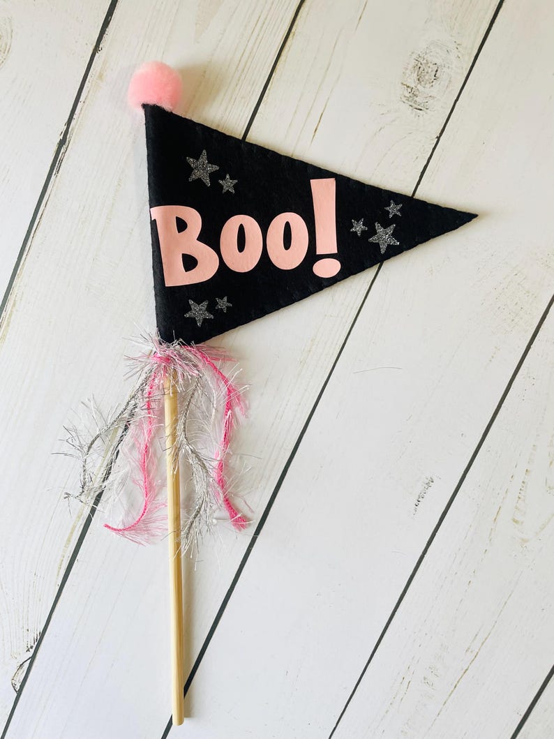 May include: A black felt flag with the word "Boo!" in pink, decorated with silver glitter stars. The flag has a pink pom-pom and is attached to a wooden dowel with pink and silver fringe, ideal for Halloween celebrations.