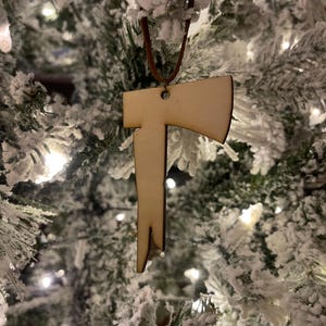 以下が含まれることがあります： 雪が積もったクリスマスツリーにぶら下がっている木製の斧のオーナメント。オーナメントは薄い茶色で、茶色の革ひもが付いています。