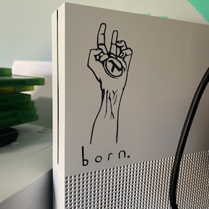 May include: A white gaming console with a black sticker of a hand holding a circle with a letter T inside. The text "born." is printed below the sticker.