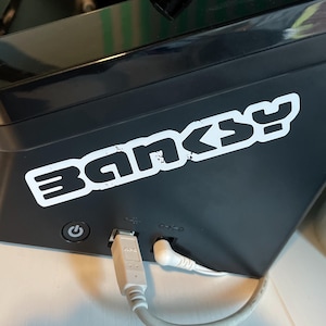 Puede incluir: Un dispositivo electrónico negro con una pegatina blanca que dice "Banksy". El dispositivo tiene un botón de encendido y dos puertos USB.
