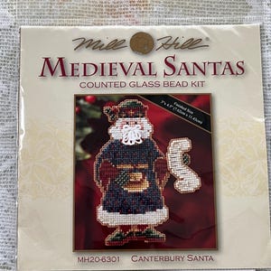 May include: A Mill Hill Medieval Santas counted glass bead kit. The kit features a cross-stitched Santa Claus design with a finished size of 3" x 4.5" (7.62cm x 11.43cm). The kit is labeled MH20-6301 Canterbury Santa.