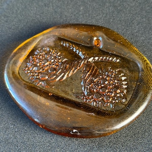 May include: An oval-shaped amber-colored glass paperweight featuring a raised, molded design of two floral clusters connected by a twisted stem. The glass has a smooth, polished surface with a translucent quality that catches the light.